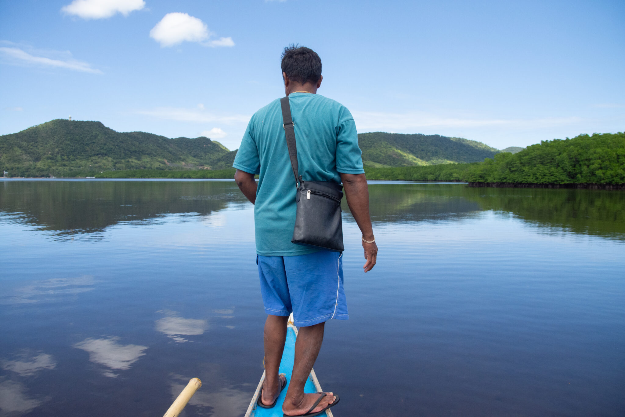 Man stands on bow of boat in shallow water, facing away from camera