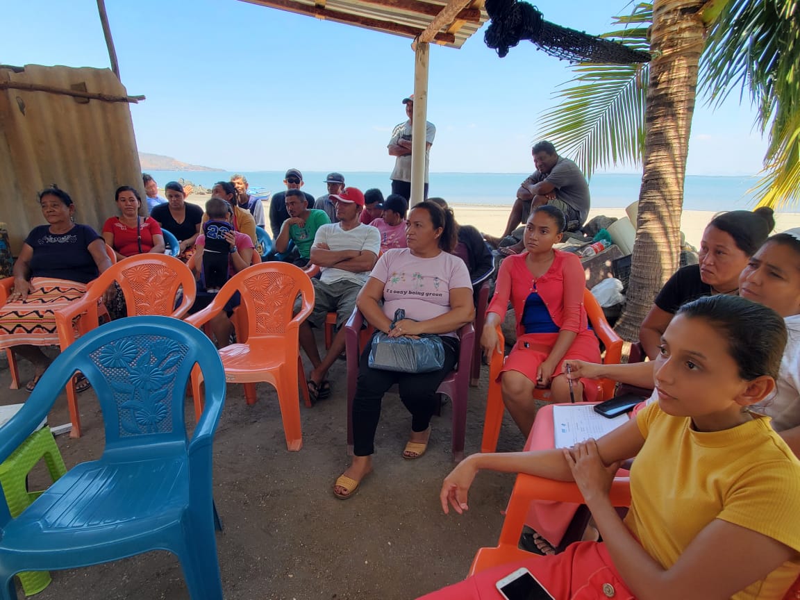 A group of people sit under a shade structure on a beach listening to a presentation