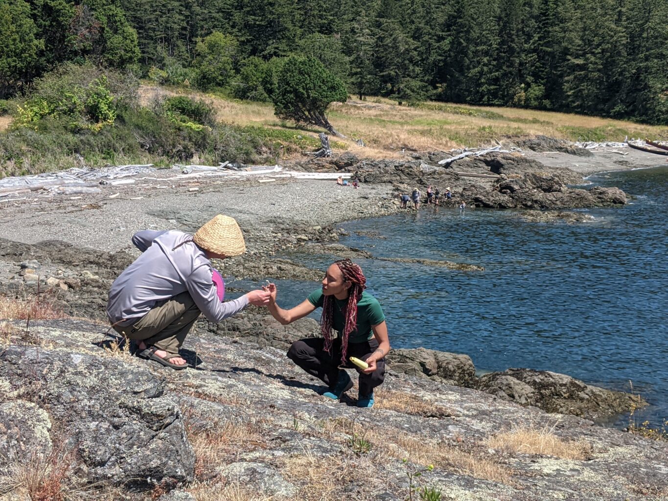 Two people crouch on the shore, looking at something in their hands