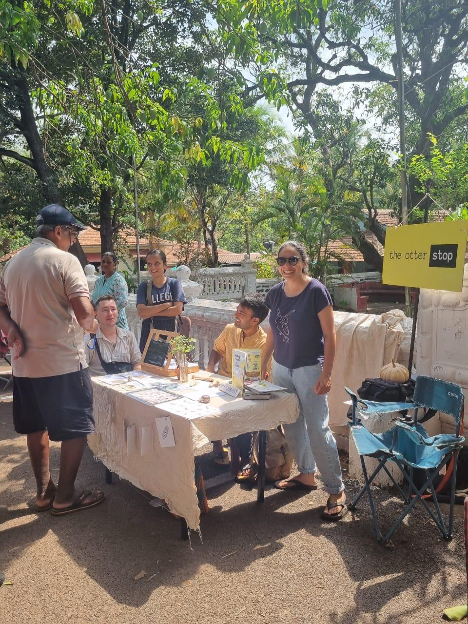 A person at a table with environmental educational materials smiles while showing it to a group of people on a sunny day