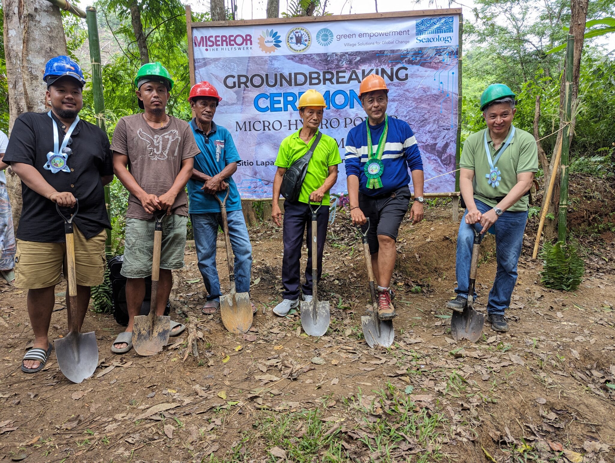 A group of people with shovels and hard hats stands in front of the groundbreaking ceremony sign