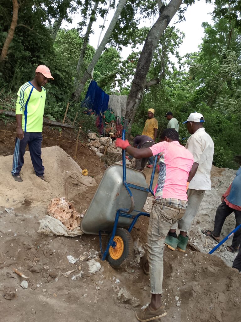 A group of people work at a construction site with a wheelbarrow, sand, and other tools